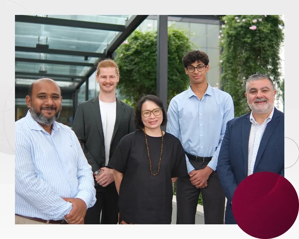 Five colleagues standing outdoors in front of office building and greenery, posing side by side.