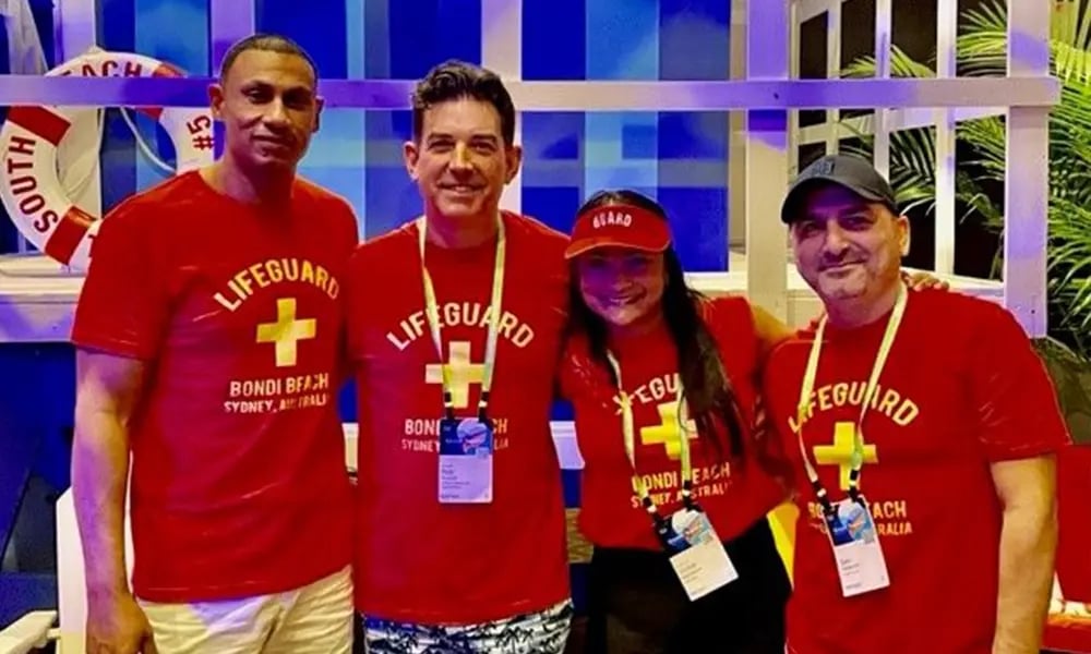 Four people wearing matching red “Lifeguard Bondi Beach” shirts pose together in a bright, beach‑themed setting.