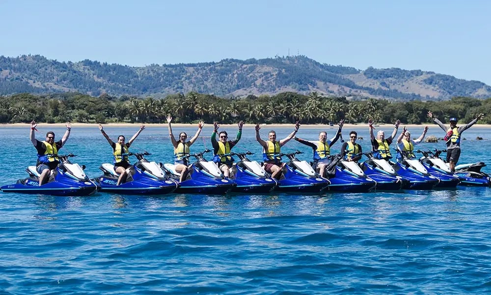 A group of people on jet skis lined up across the water with their arms raised, enjoying a team activity against a scenic coastal backdrop.