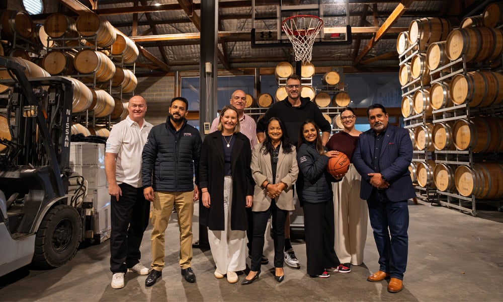Group of people standing in a warehouse with stacked wine barrels and a basketball hoop.