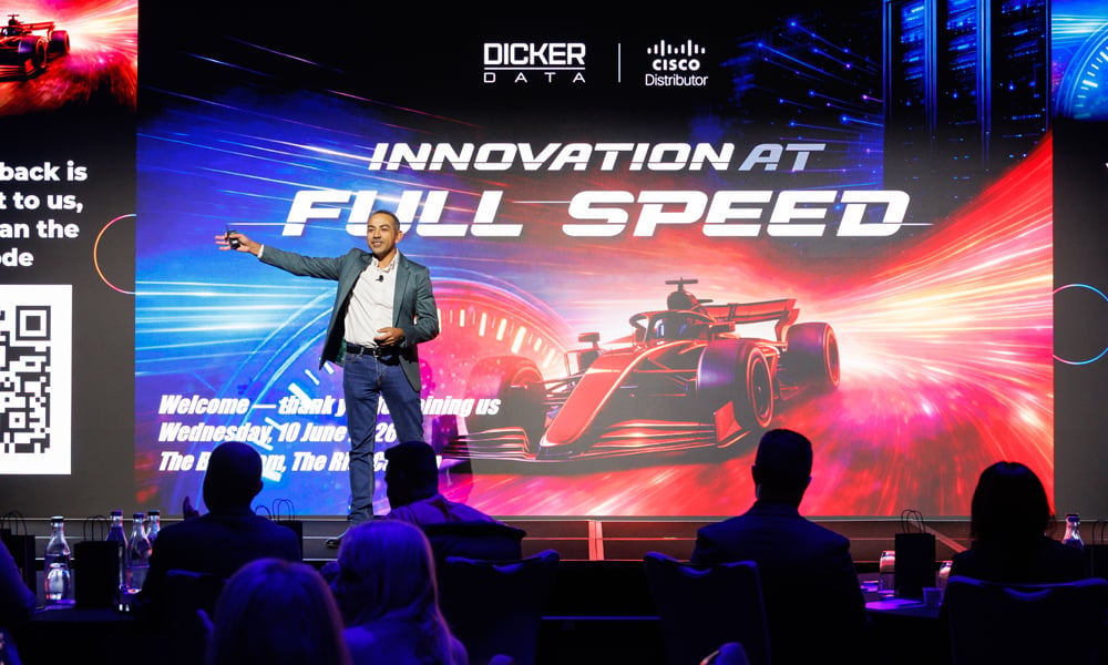 Presenter on stage at event with “Innovation at Full Speed” screen and race car graphics.