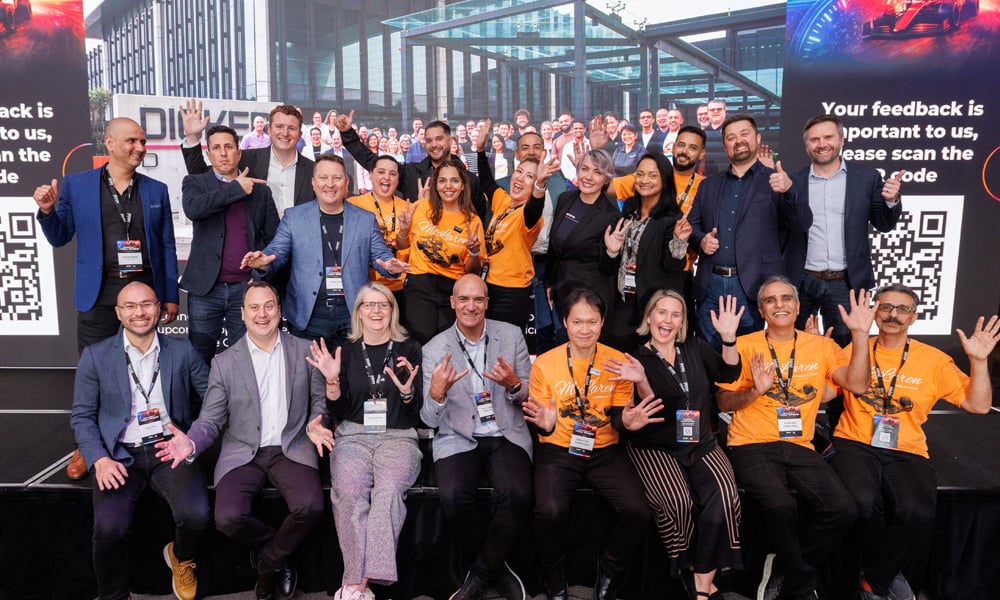 Large group on stage at event wearing orange shirts, posing in front of branded backdrop with QR codes.