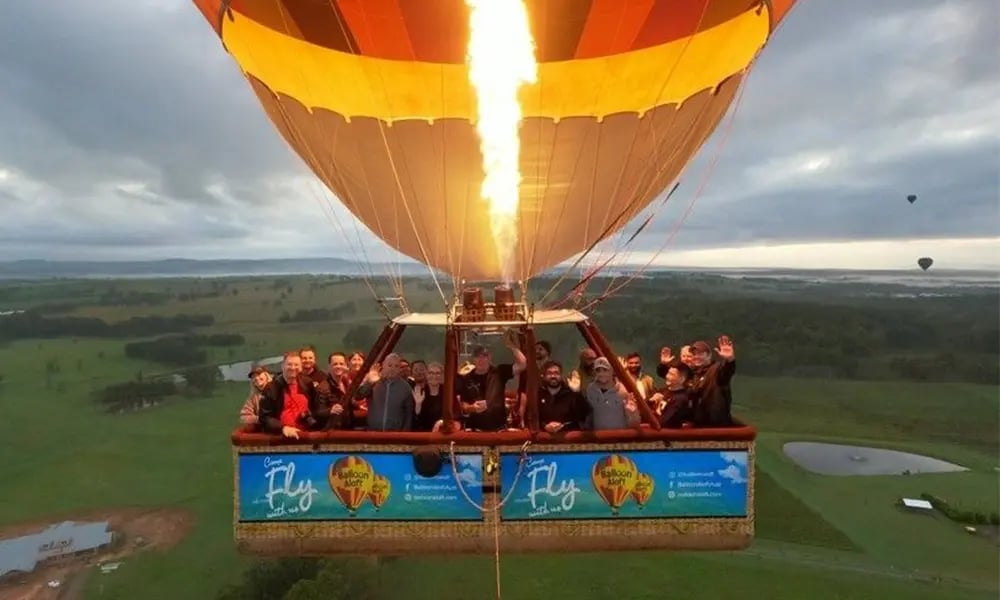 A large group stands inside a hot‑air balloon basket mid‑flight, floating above green fields at sunrise.