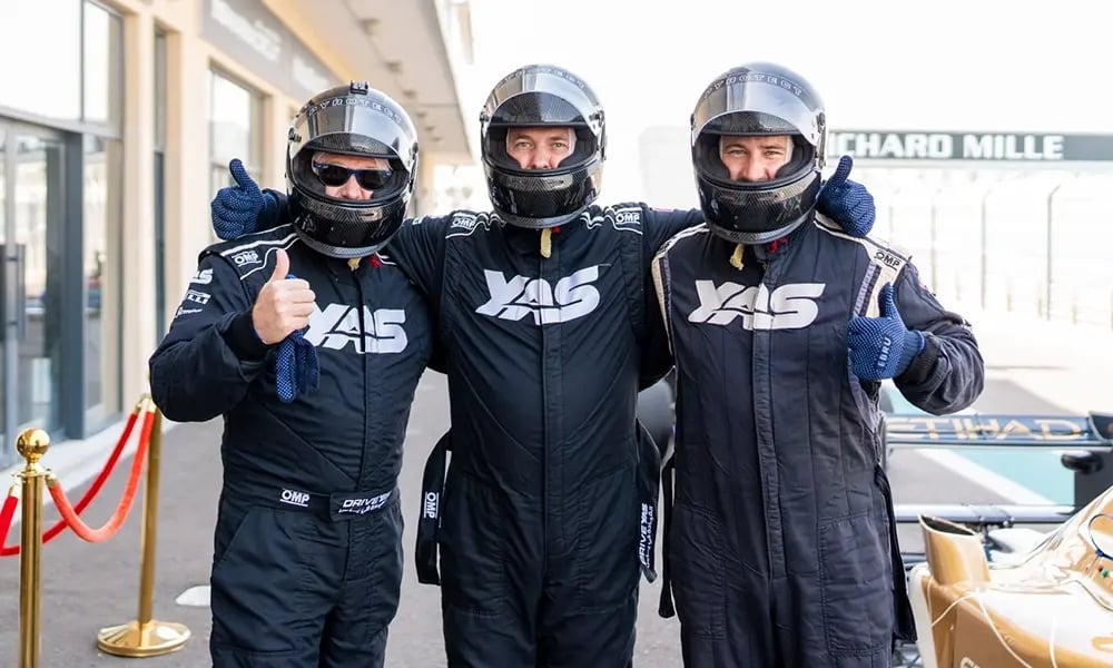 Three people in racing suits and helmets standing together with thumbs up.