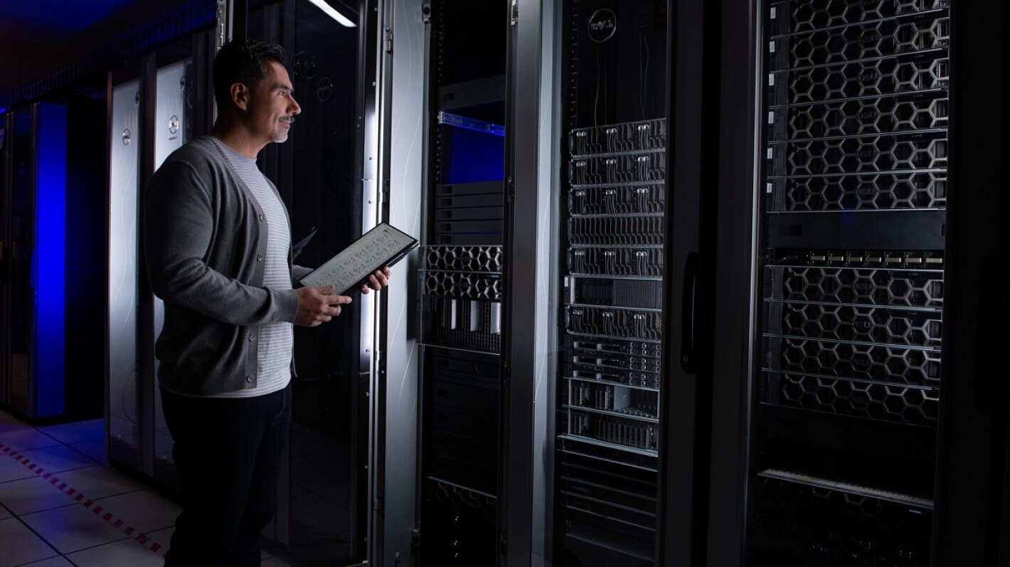 Person using a tablet while standing in a data centre aisle with server racks, representing AI or IT infrastructure management.