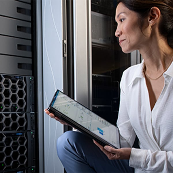 Person kneeling beside server racks in a data centre, holding a tablet and reviewing system or network information.