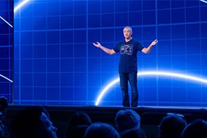 Speaker standing on a stage in front of a blue digital backdrop, addressing an audience.
