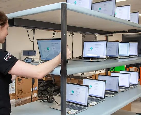 Person reaching toward a row of powered-on laptops on multi-level racks in a device testing or setup lab