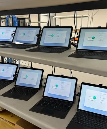 Rows of laptops on multi-tier racks in a device setup lab, each showing a configuration screen