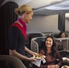 Flight attendant in uniform serves a smiling passenger a meal in a luxurious airplane cabin. The atmosphere appears welcoming and comfortable.