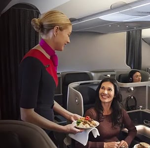 Flight attendant in uniform serves a smiling passenger a meal in a luxurious airplane cabin. The atmosphere appears welcoming and comfortable.