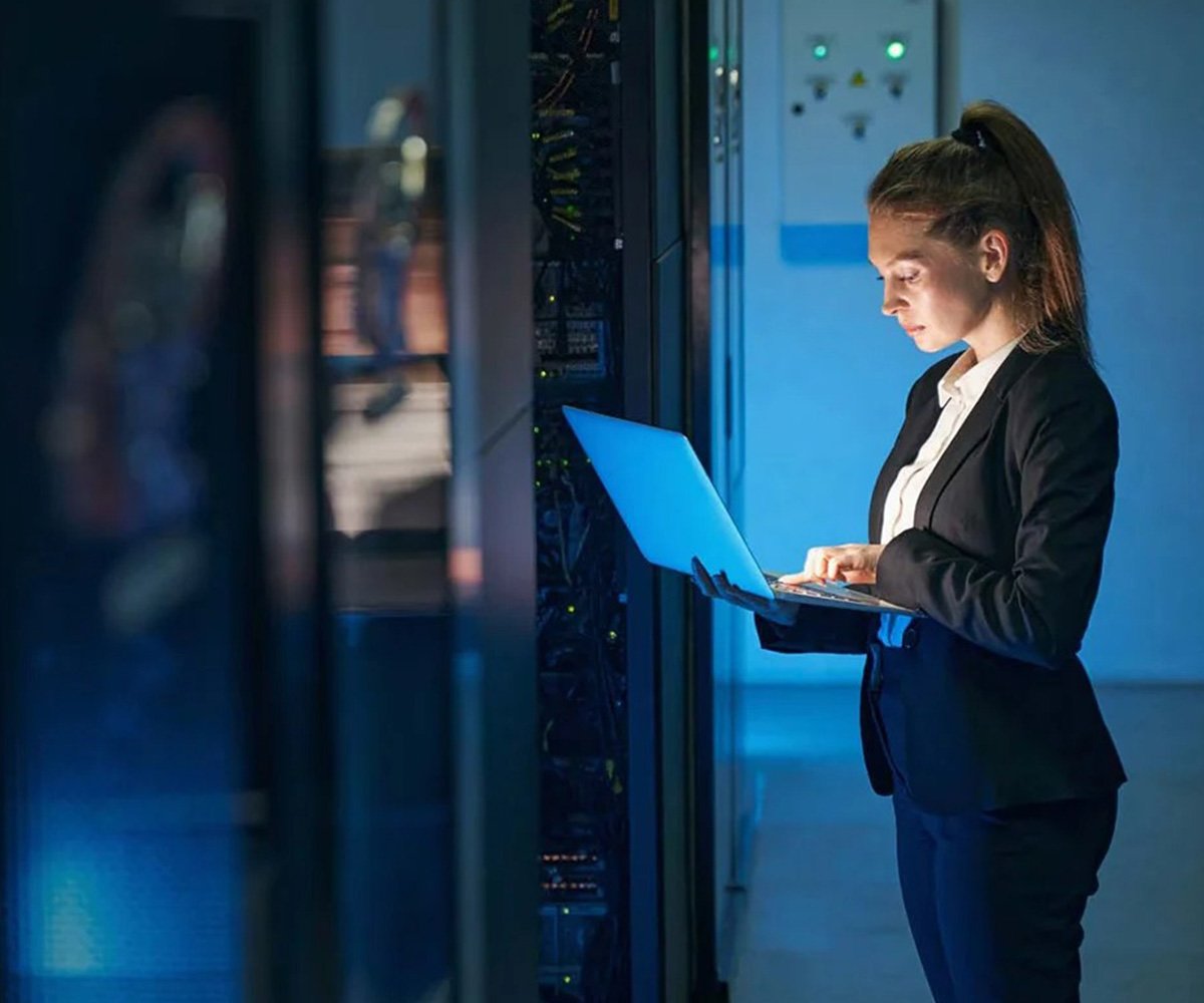 Person using a laptop in a server room, standing beside a rack of network equipment under cool blue lighting