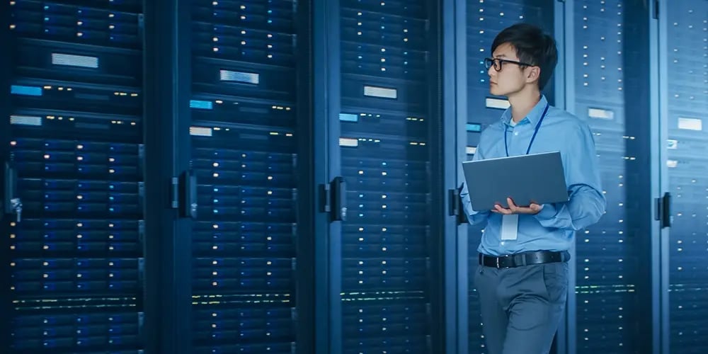 Person holding a laptop while standing in a server room with rows of illuminated server racks