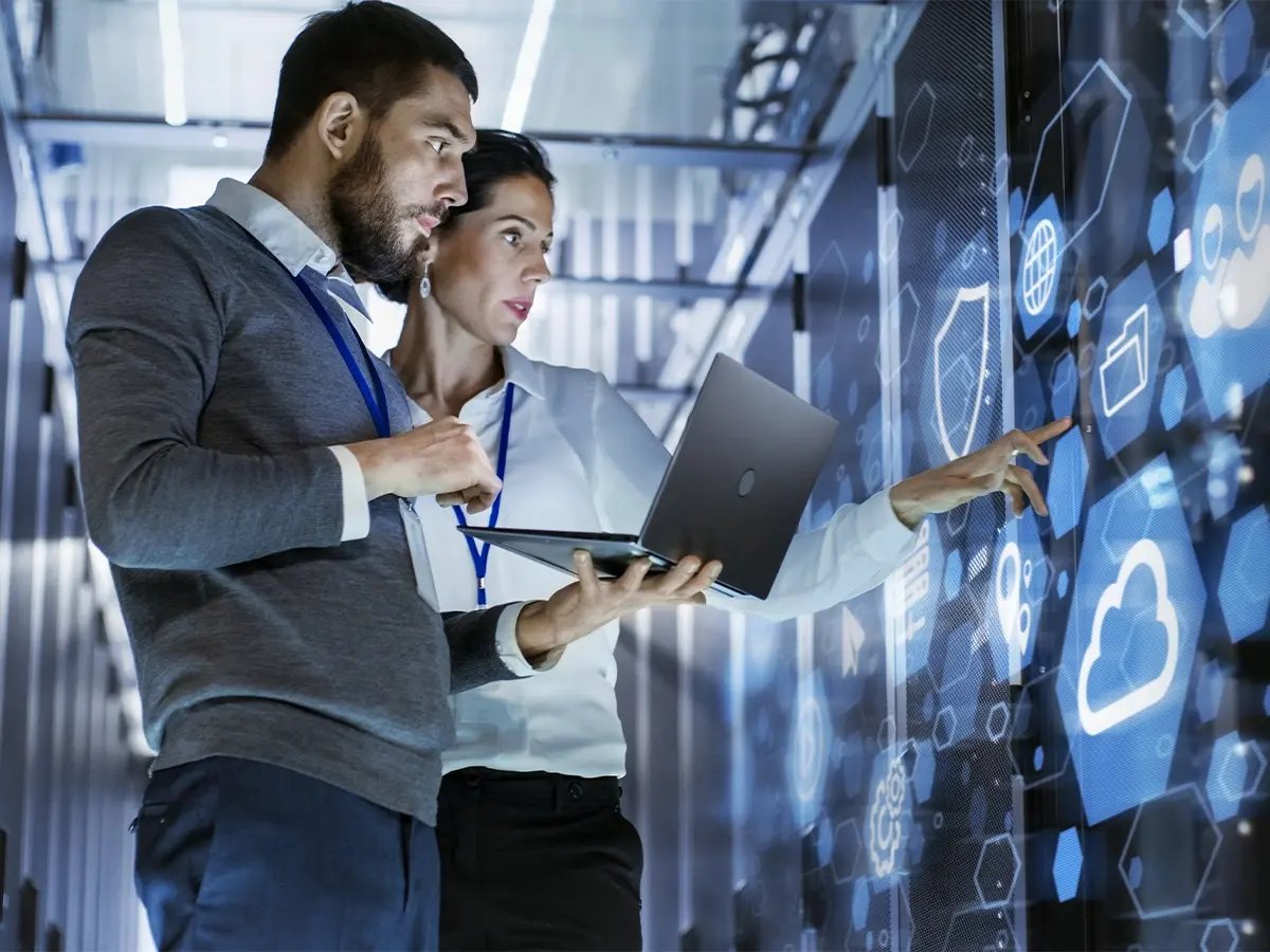 Two people in a server room reviewing data on a laptop and pointing at a digital wall display with cloud and network icons.
