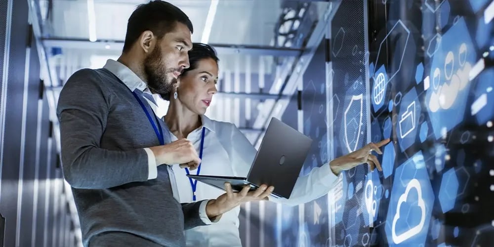 Two people in a server room reviewing data on a laptop and pointing at a digital wall display with cloud and network icons.