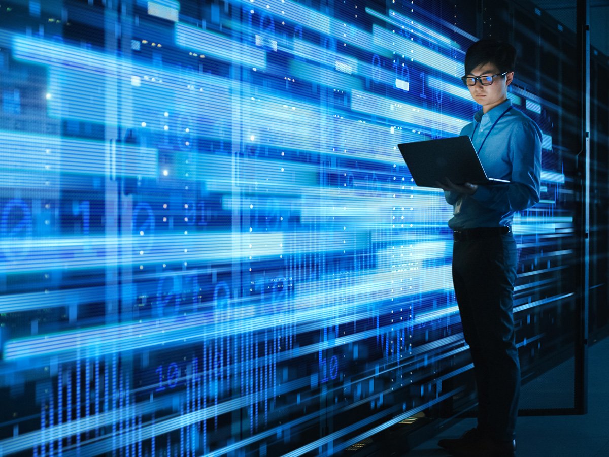 Person holding a laptop while standing in front of illuminated server racks with blue light effects.