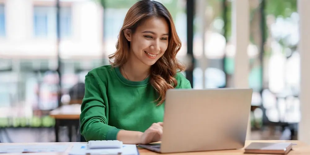 Person in a green sweater working on a laptop at a wooden table with papers and a pen nearby.