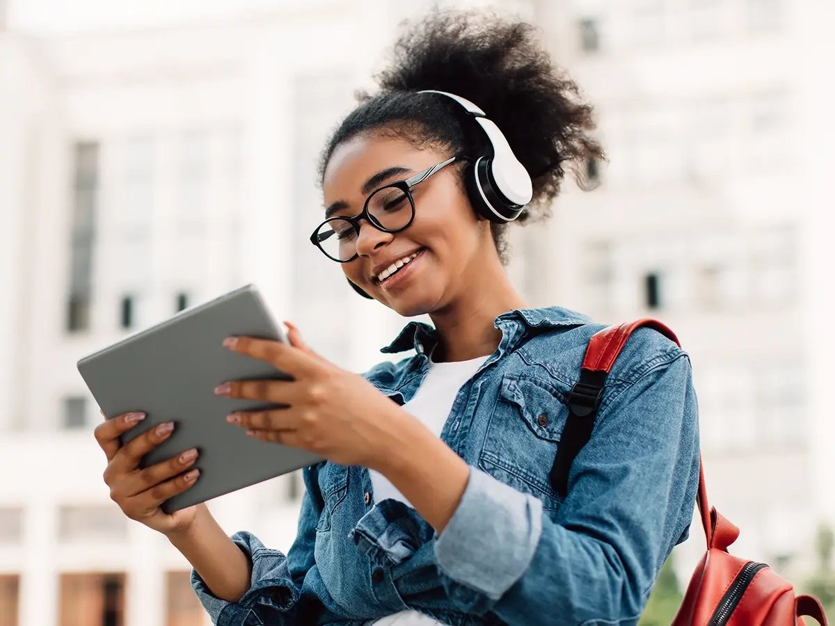 Person wearing headphones and carrying a red backpack while holding a tablet outdoors.
