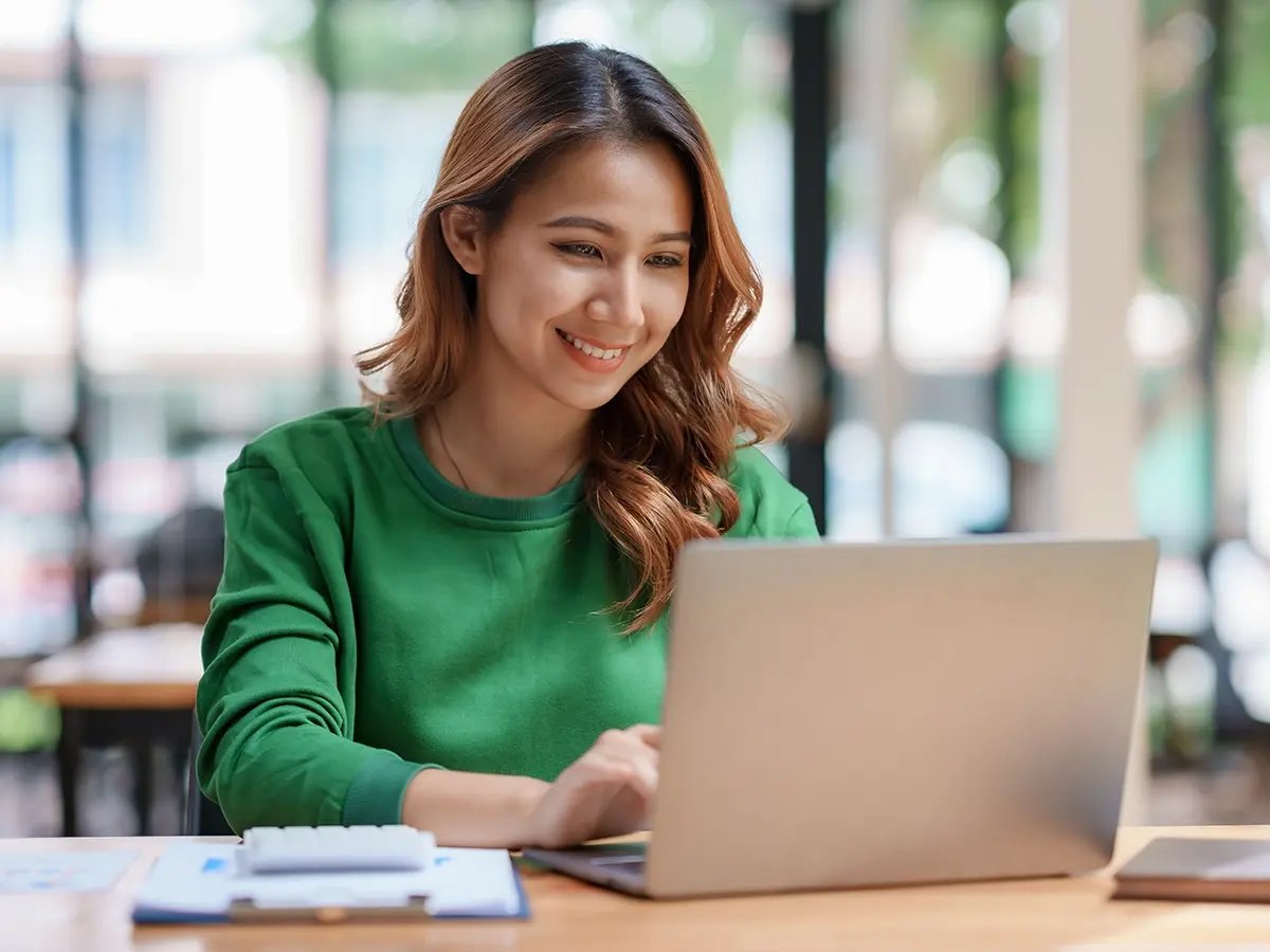 Person in a green sweater working on a laptop at a wooden table with papers and a pen nearby.