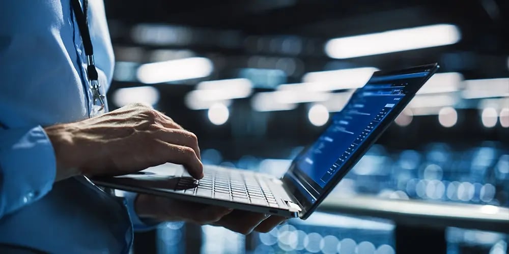 Hands typing on a laptop in a server room.