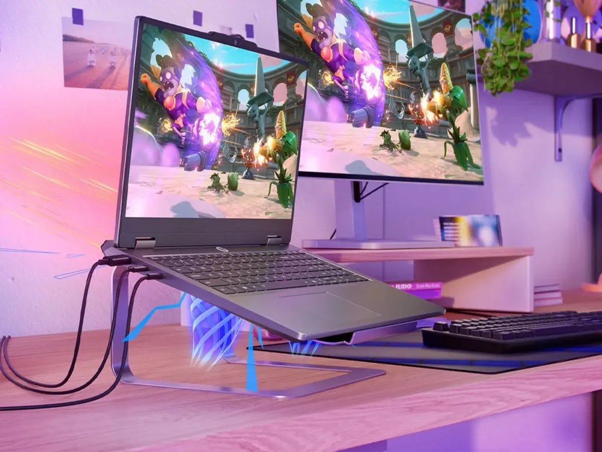 Gaming laptop on a stand connected to external monitor, both displaying a colorful action game, with keyboard and accessories on a desk.