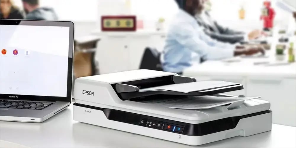 Epson document scanner placed next to an open laptop on a white desk in an office setting.