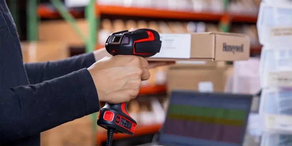 Person using a handheld barcode scanner to scan a box in a warehouse setting.