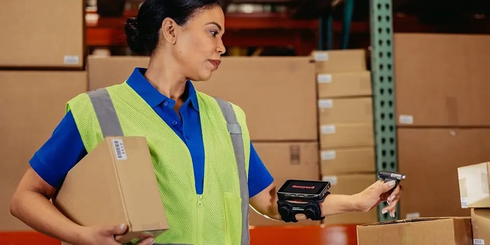 Warehouse worker in a safety vest holding a box and using a wearable barcode scanner.