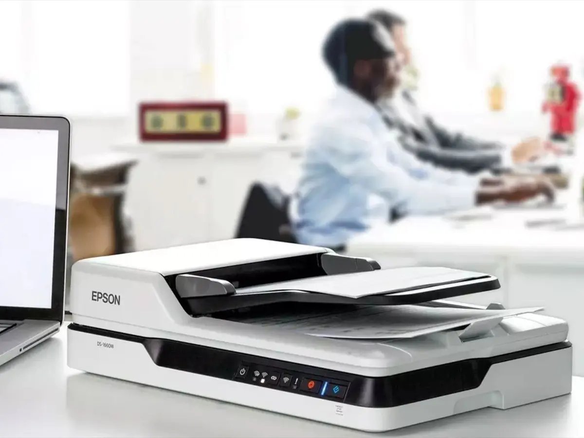 Epson document scanner placed next to an open laptop on a white desk in an office setting.