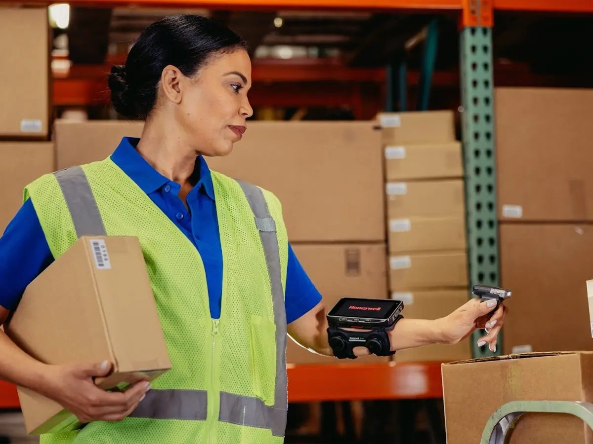Warehouse worker in a safety vest holding a box and using a wearable barcode scanner.