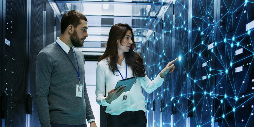 Two people in a server room reviewing data on a tablet and pointing at a network panel.