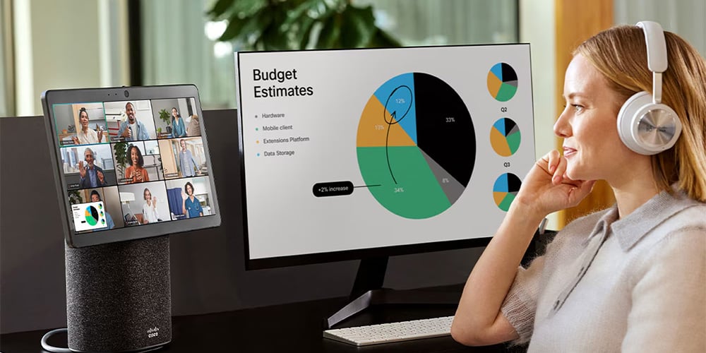 Person at desk with headphones, video call on one screen and budget charts on another.