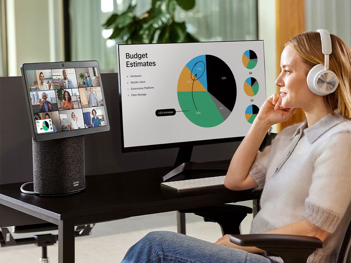 Person at desk with headphones, video call on one screen and budget charts on another.