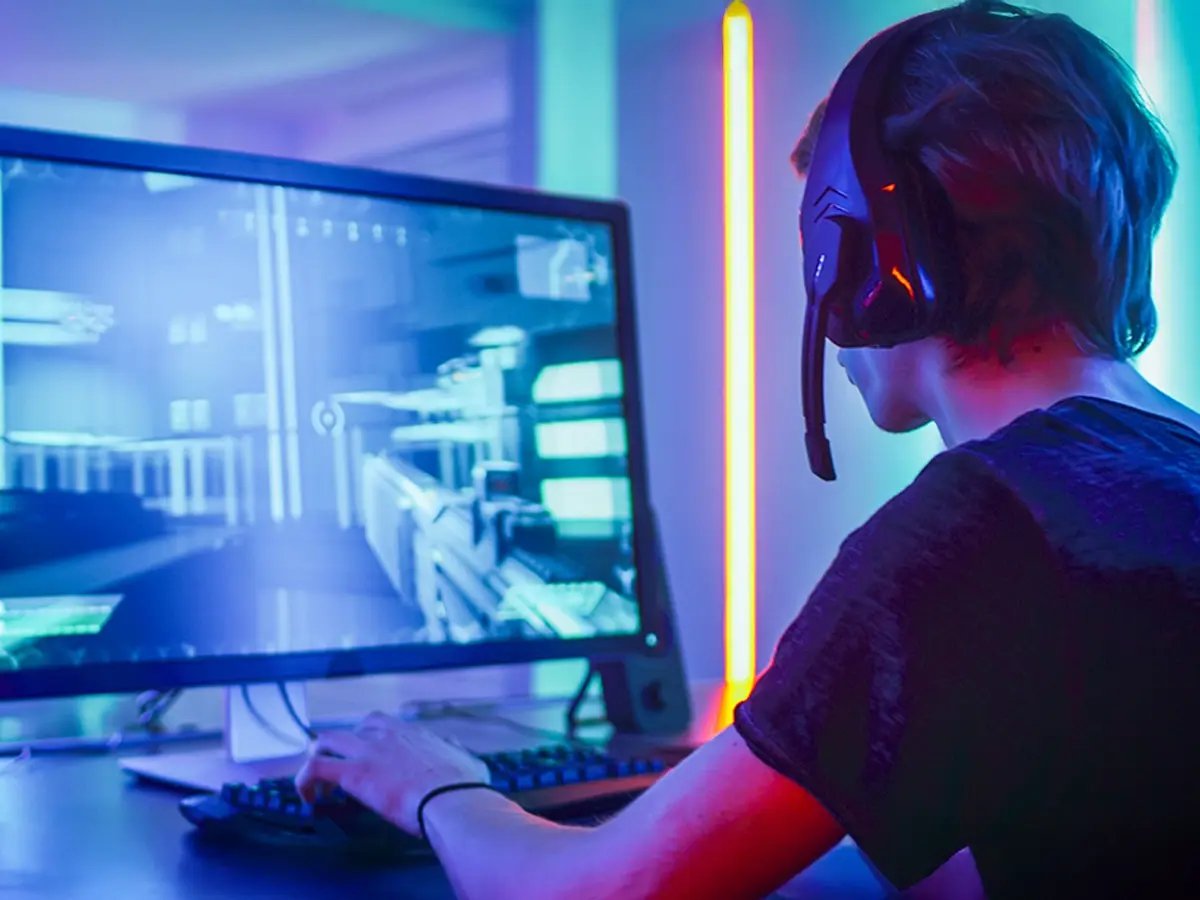 A person wearing a headset plays a video game on a computer, surrounded by neon lights. The atmosphere is immersive and focused, with vibrant colors.