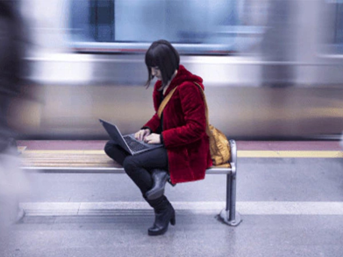 Individual using a laptop while seated on a bench at a train station.