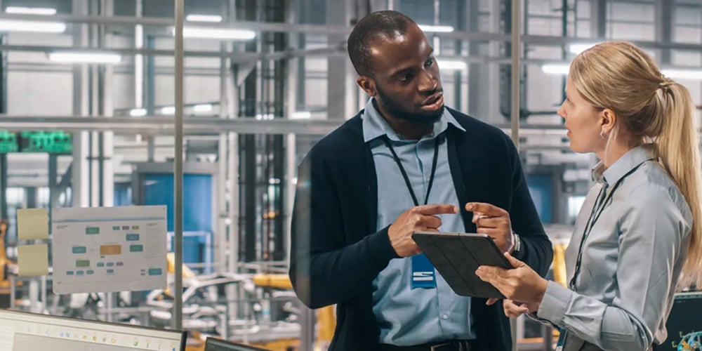 Two people standing in a factory office reviewing data on a tablet with monitors nearby.