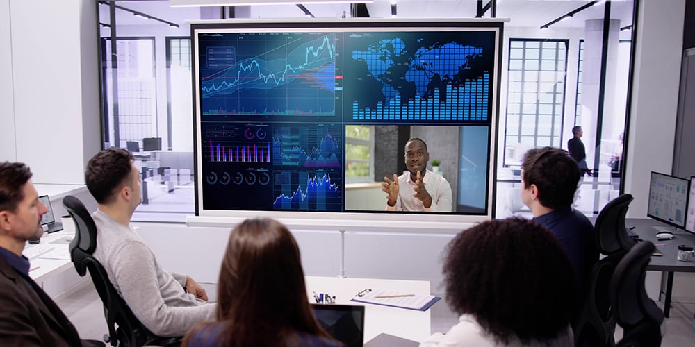 Group in a meeting room viewing large screens displaying global charts, graphs, and a video call.