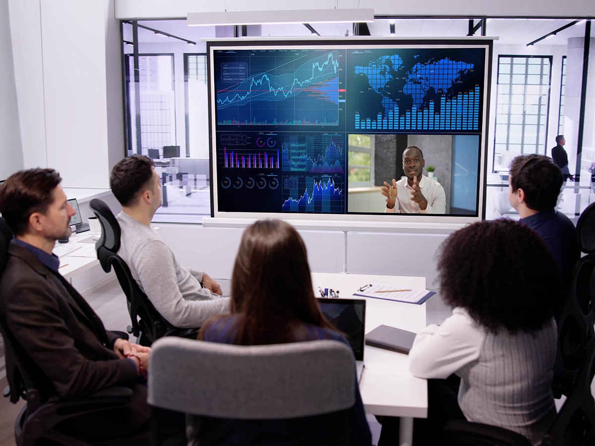 Group in a meeting room viewing large screens displaying global charts, graphs, and a video call.