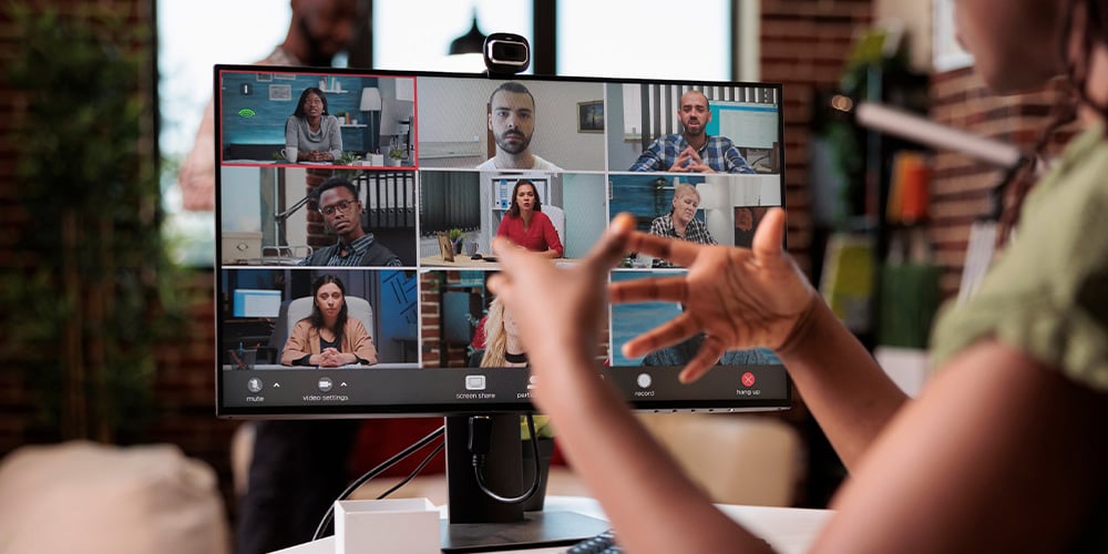 Person at a desk speaking during a video call displayed on a monitor with multiple participants.