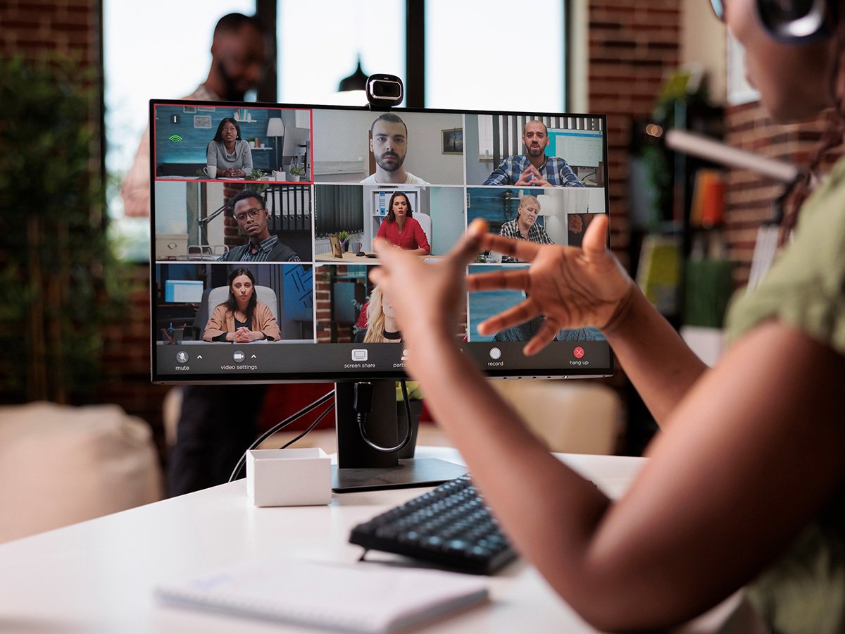 Person at a desk speaking during a video call displayed on a monitor with multiple participants.