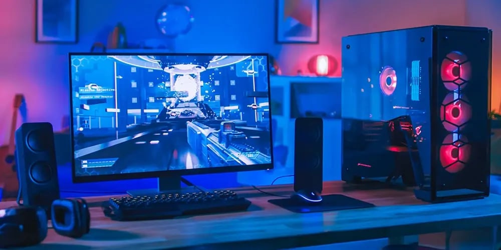 Desktop gaming setup with a monitor displaying a first‑person game, illuminated PC tower, keyboard, and speakers.