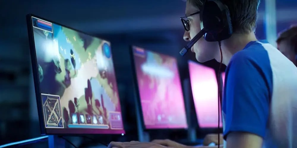 Person wearing a headset playing a video game at a desk with multiple monitors, keyboard, and mouse in a dimly lit room.