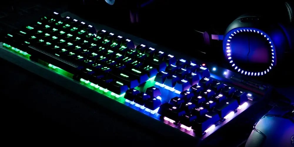 RGB‑lit mechanical keyboard on a dark desk, glowing green, blue, and purple, with a gaming headset nearby.