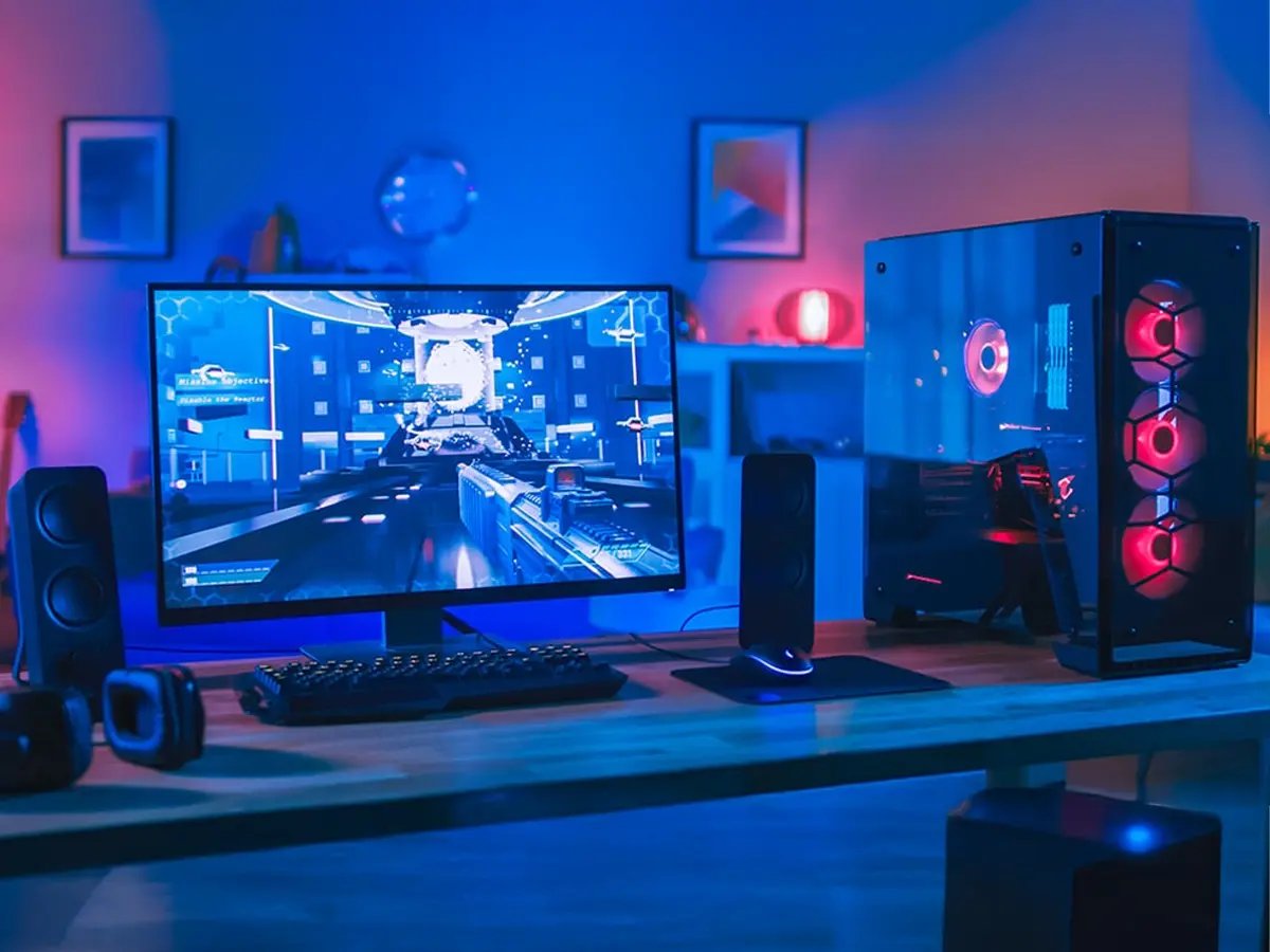 Desktop gaming setup with a monitor displaying a first‑person game, illuminated PC tower, keyboard, and speakers.