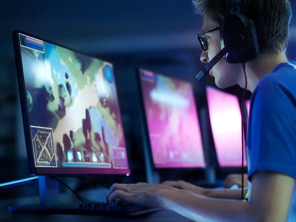 Person wearing a headset playing a video game at a desk with multiple monitors, keyboard, and mouse in a dimly lit room.