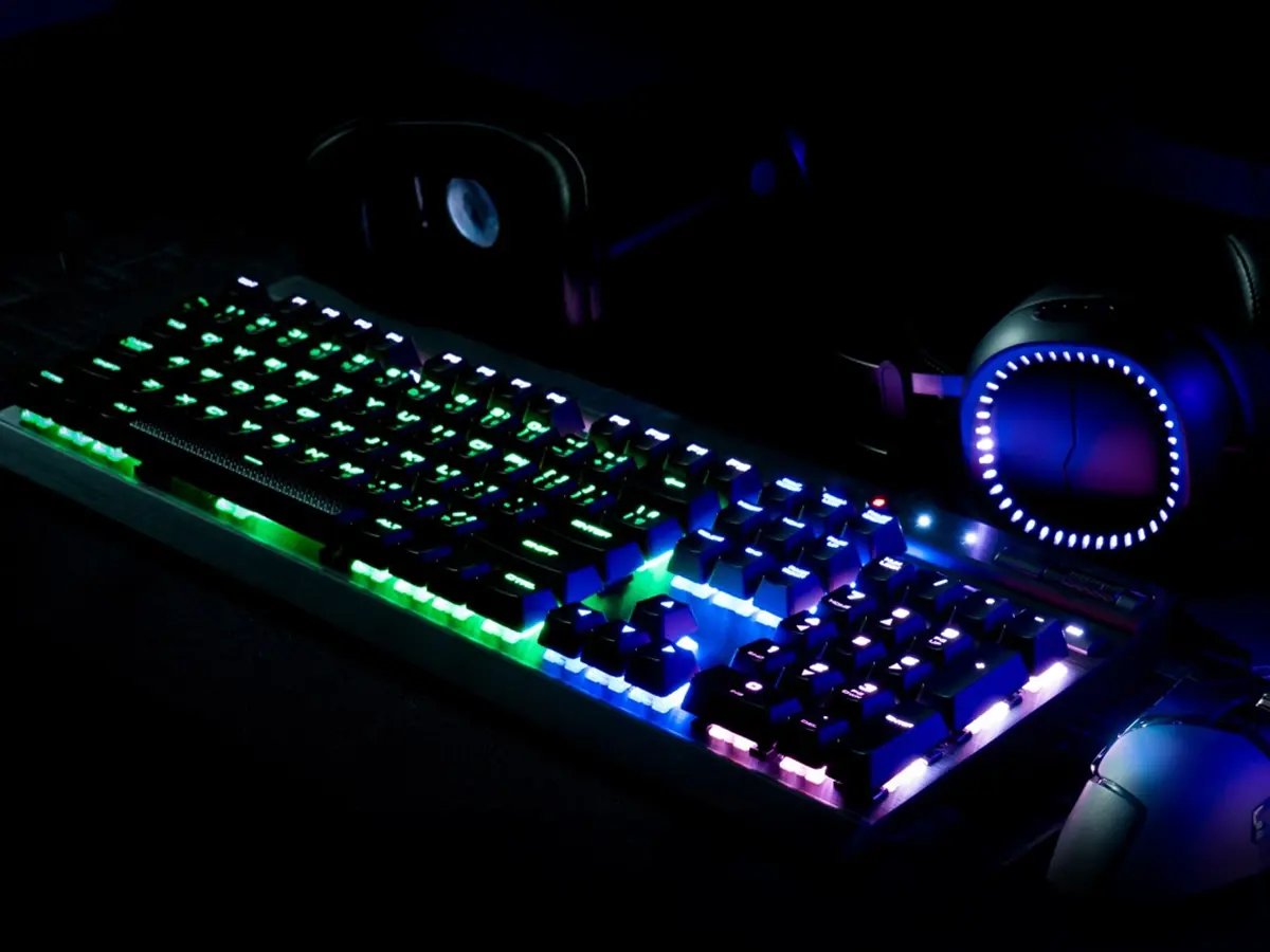 RGB‑lit mechanical keyboard on a dark desk, glowing green, blue, and purple, with a gaming headset nearby.