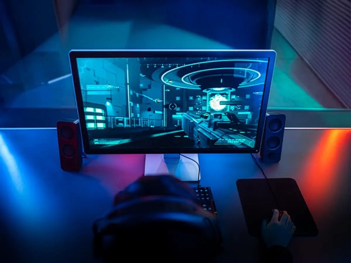 Overhead view of a desktop monitor showing a first‑person video game, with keyboard, mouse, speakers, and blue lighting.