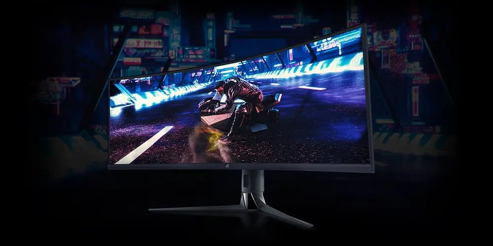 A large curved monitor displays a futuristic racing game. A motorcyclist speeds through a neon-lit city. A keyboard and mouse are in front, set against a dark blue backdrop.