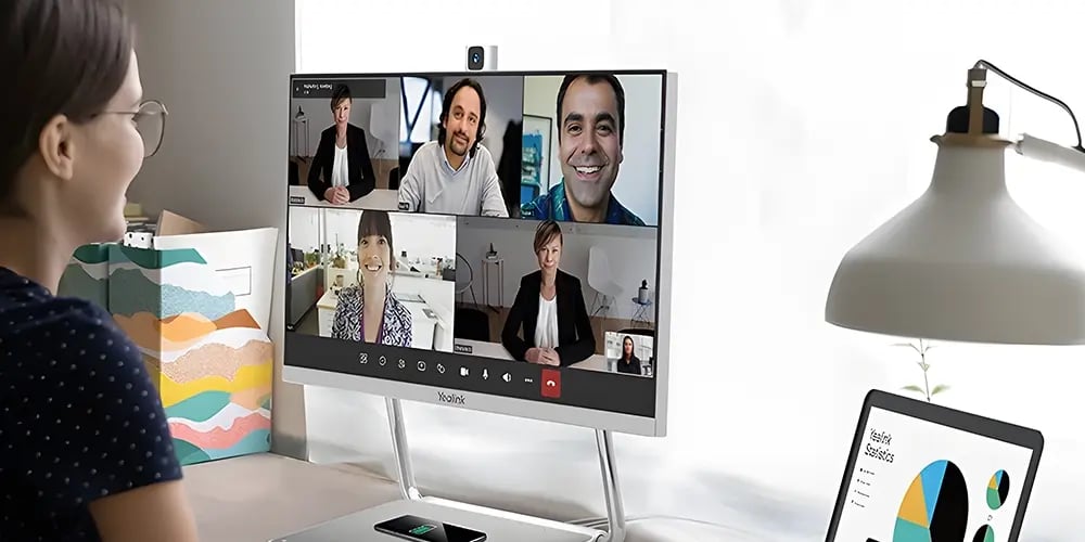 Person seated at a desk participating in a video call displayed on a large monitor in a home office.