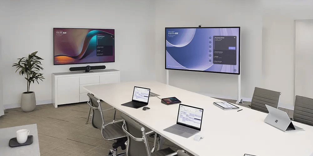 Modern meeting room with a long table, chairs and two large wall‑mounted displays showing presentation content.